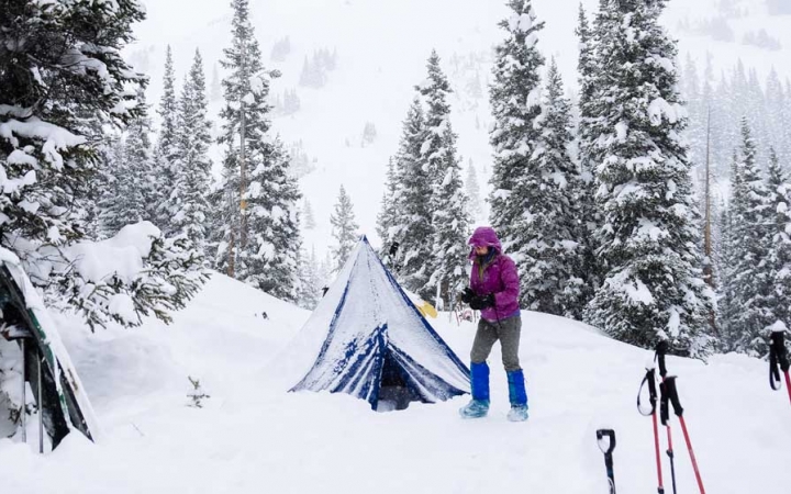 a person stands outside a snow-dusted tent. there are trekking poles in the foreground, and trees in the background. 